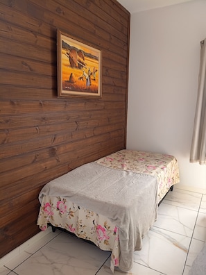 Una habitación con pared de madera, una cama con colcha estampada y un cuadro de una escena campestre.