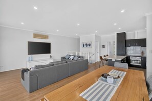 A modern living room with a grey sofa, a wooden dining table, and a kitchen area with dark cabinets and a stainless steel oven. - 9 Barbara Street, Port Elliot Near Horseshoe Bay & Greenbay (Port Elliot)