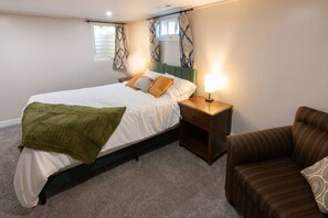 A bedroom with a bed, bedside table, lamp, and a striped armchair. - Deco Garden Lounge - With Sauna Access! (Ogden)