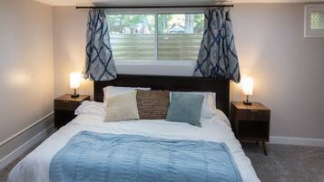 Ein Schlafzimmer mit einem großen Bett, zwei Nachttischen mit Lampen, einem Fenster mit gemusterten Vorhängen und Blick auf eine grüne Landschaft.