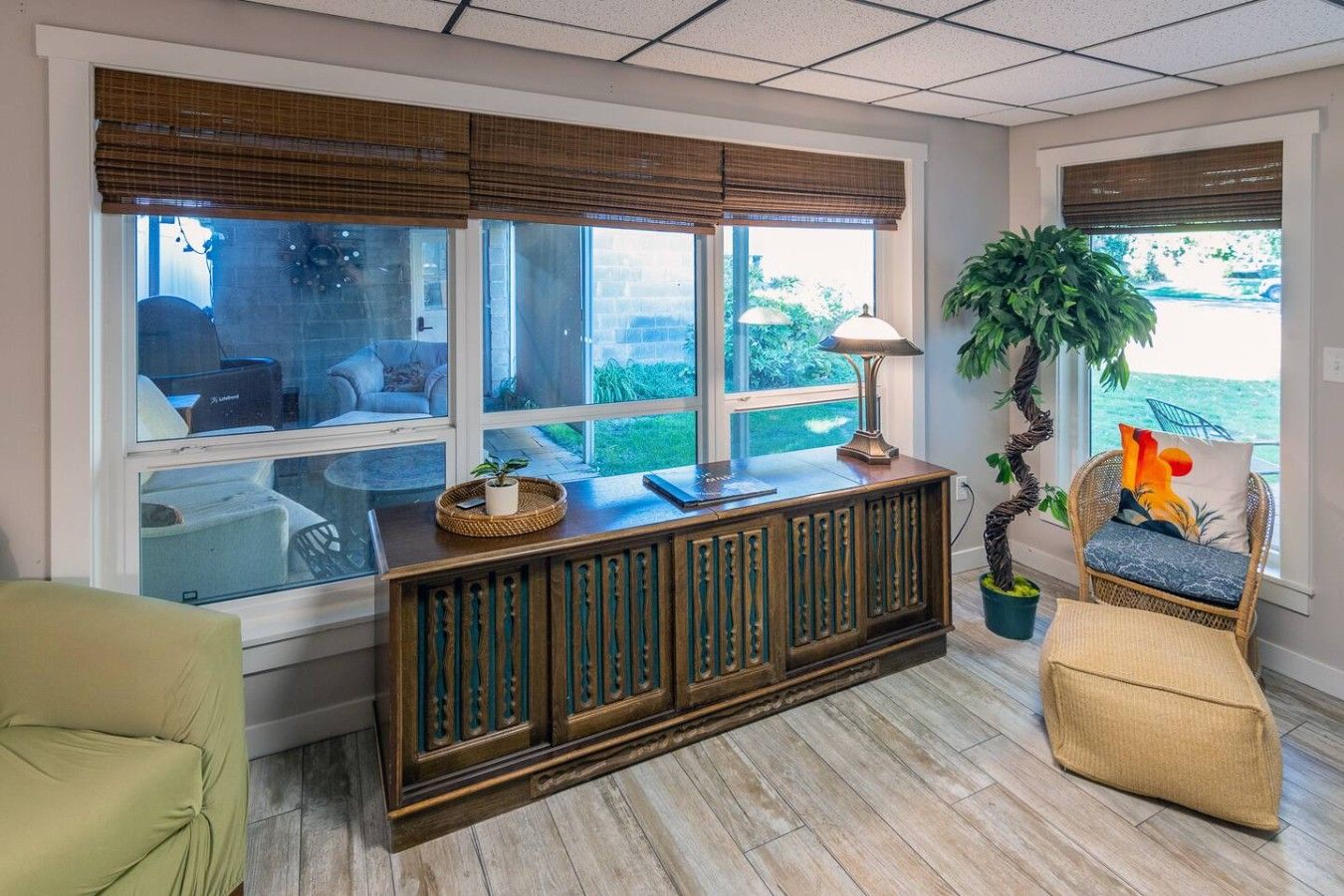 A room with a wooden console table, a lamp, a potted plant, a wicker chair, and a view of the outdoors through large windows.