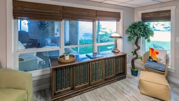 A room with a wooden console table, a lamp, a potted plant, a wicker chair, and a view of the outdoors through large windows.