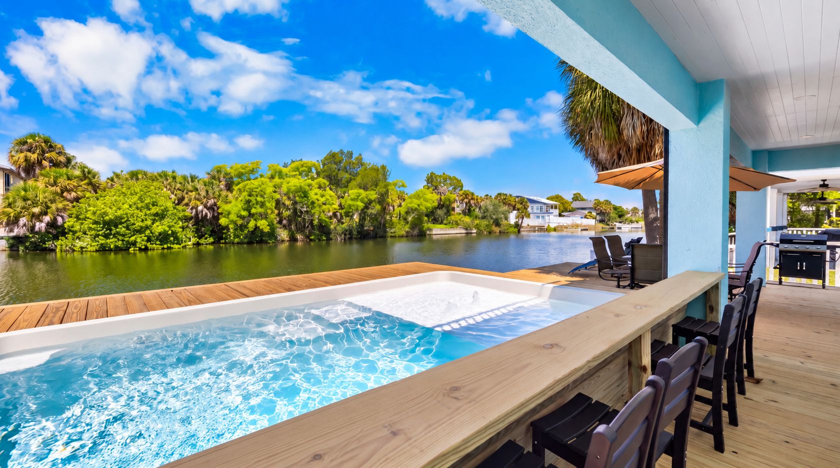 A pool area with a wooden deck, lounge chairs, and a view of a body of water and greenery.