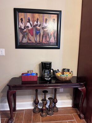 A dining table with a coffee maker, a basket of snacks, and a framed painting on the wall. - The Mcdonough Luxury Island Suite (McDonough)