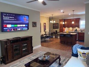 A living room with a flat-screen TV displaying a documentary, a coffee table with decorative items, a dining area with chairs, and a kitchen with wooden cabinets and a bar. - The Mcdonough Luxury Island Suite (McDonough)