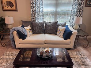 Un salon avec un canapé beige, des coussins à motifs, une table basse en verre agrémentée d’objets décoratifs et des rideaux à motifs.