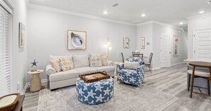 A modern living room with a sofa, ottoman, and chairs, a dining table, and a hallway with doors. - 3BR Coastal Home in Lost Bay (Orange Beach)