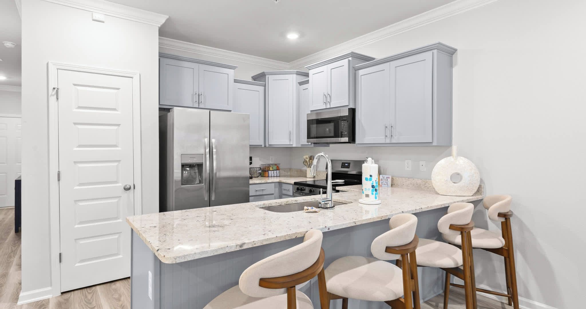 A modern kitchen with a stainless steel refrigerator, a microwave, and a granite countertop. There are three bar stools and a sink with a faucet.