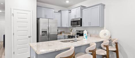 A modern kitchen with a stainless steel refrigerator, a microwave, and a granite countertop. There are three bar stools and a sink with a faucet.