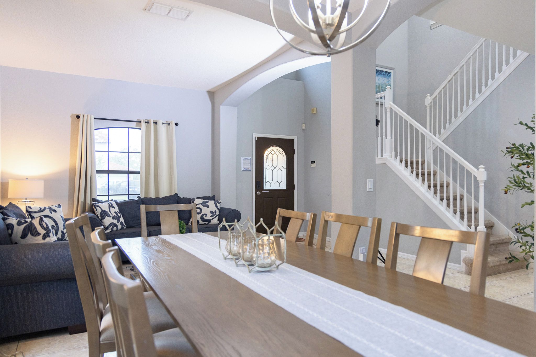 A dining area with a wooden table, chairs, and candle holders. There is a staircase to the right and a door with a stained glass window in the background.