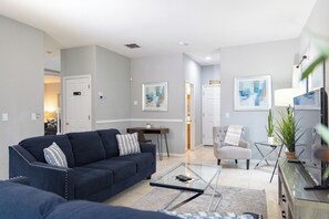 Ein modernes Wohnzimmer mit einem dunkelblauen Sofa, einem Glastisch und einem grauen Sessel. Es gibt einen Essbereich mit einem kleinen Tisch und Stühlen sowie einen Fernseher auf einem Lowboard.