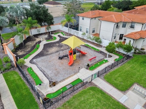 Ein Spielplatz mit Rutsche und Klettergerüsten, umgeben von einem eingezäunten Bereich mit grüner Wiese und einem befestigten Weg.