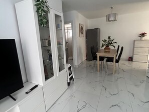 Een moderne woonkamer met een flatscreen televisie, een eettafel met stoelen, een witte kast met glazen deuren en een potplant.
