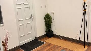 Una habitación con suelo de madera, un tapete gris, una puerta blanca, una planta en maceta, una lámpara de pie y un felpudo negro.
