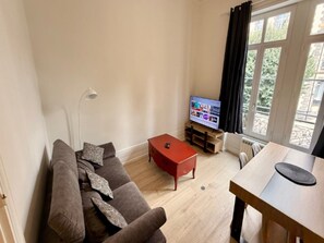 A living room with a sofa, a coffee table, a TV on a wooden stand, and a window with curtains. - Apartment Near Parc Fenestre (La Bourboule)