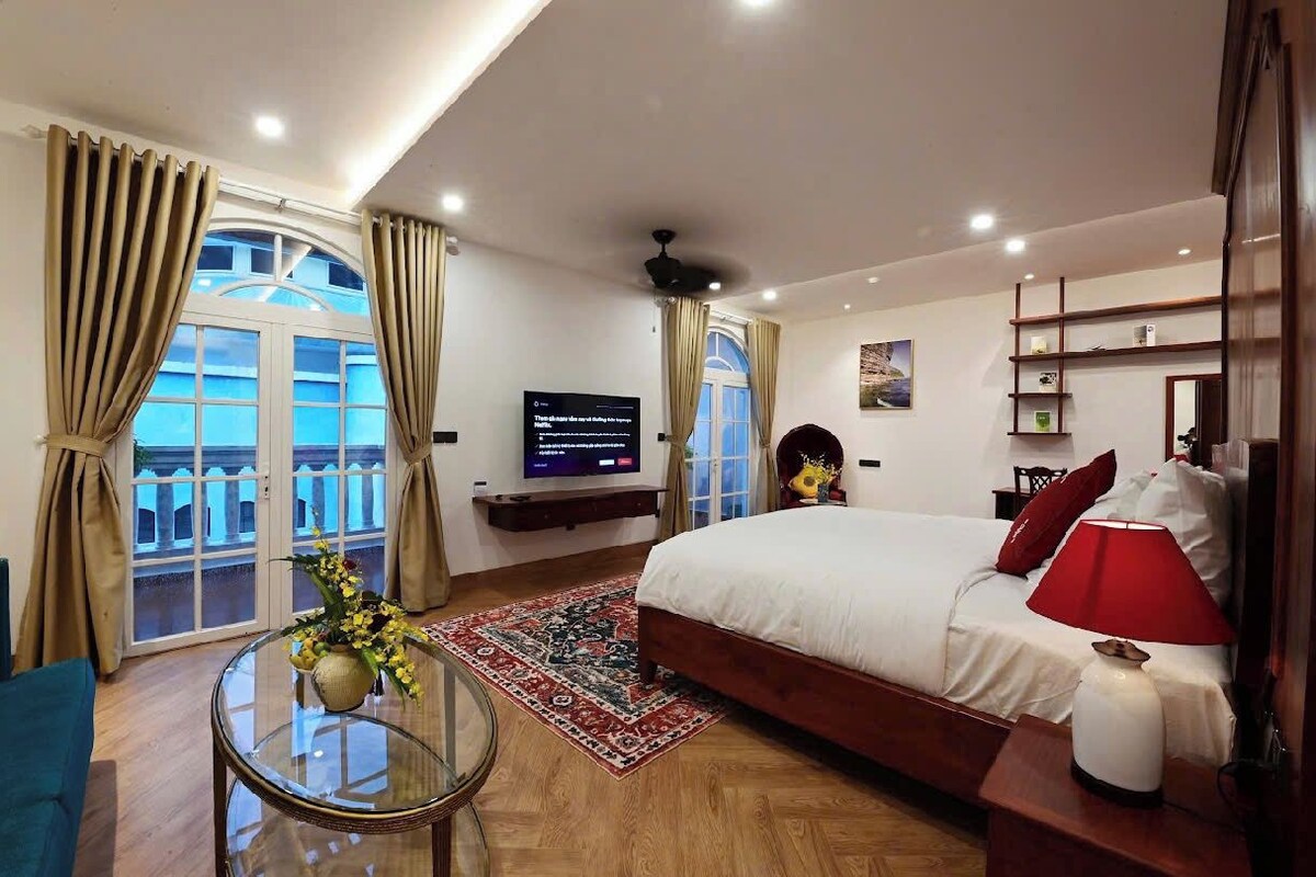 Een hotelkamer met een groot bed, een televisie, een balkon met gordijnen, een tafeltje met een vaas en een rode lamp.