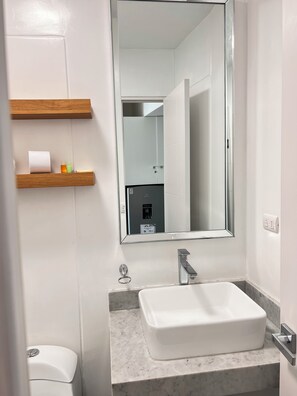 A bathroom with a white sink, a mirror, and a toilet. There are wooden shelves above the sink with toiletries. - Vrbo Property (Barranco)