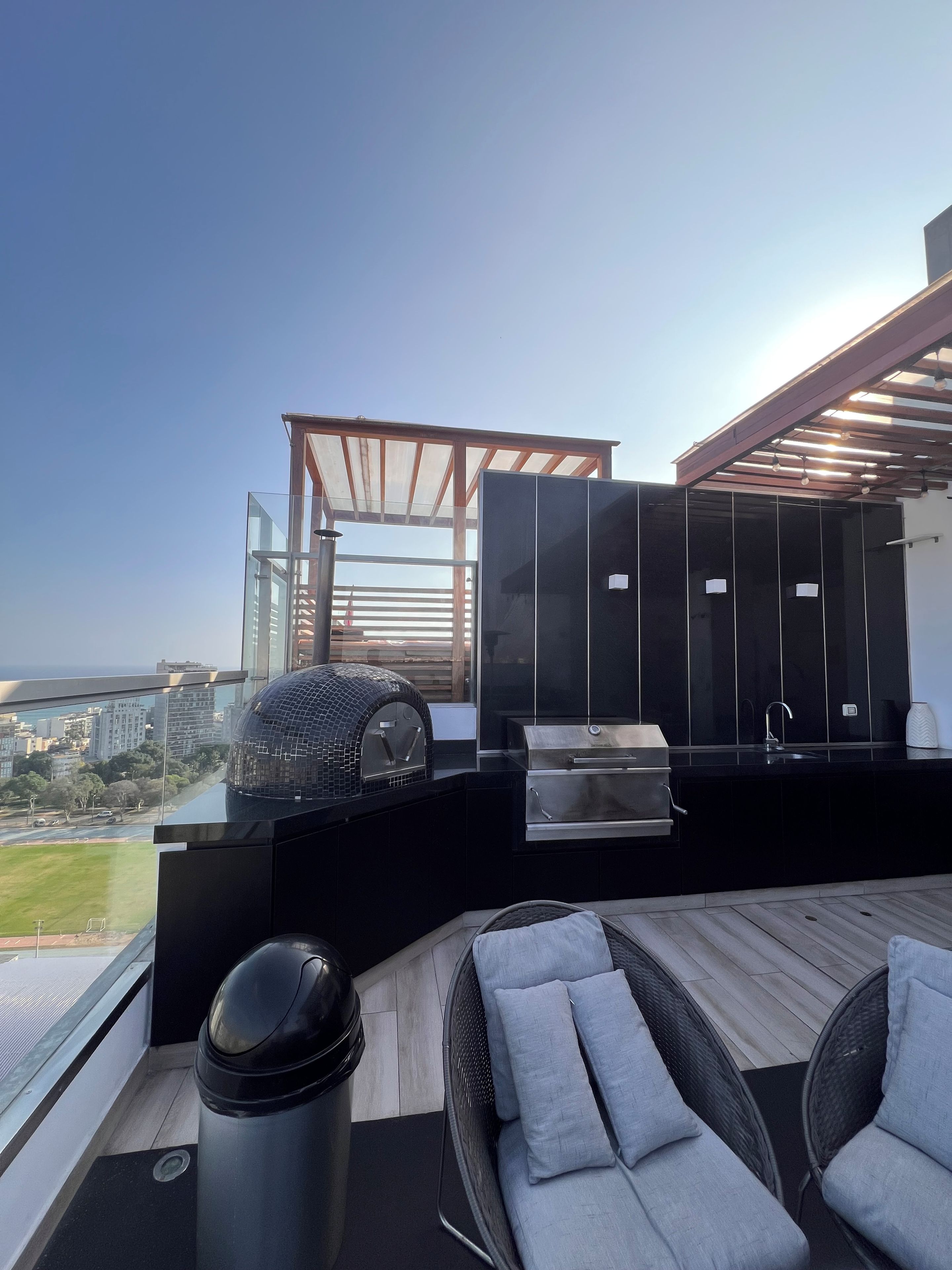 Une cuisine extérieure moderne, dotée d’un barbecue, d’un espace détente avec des coussins et offrant une vue sur le paysage urbain.