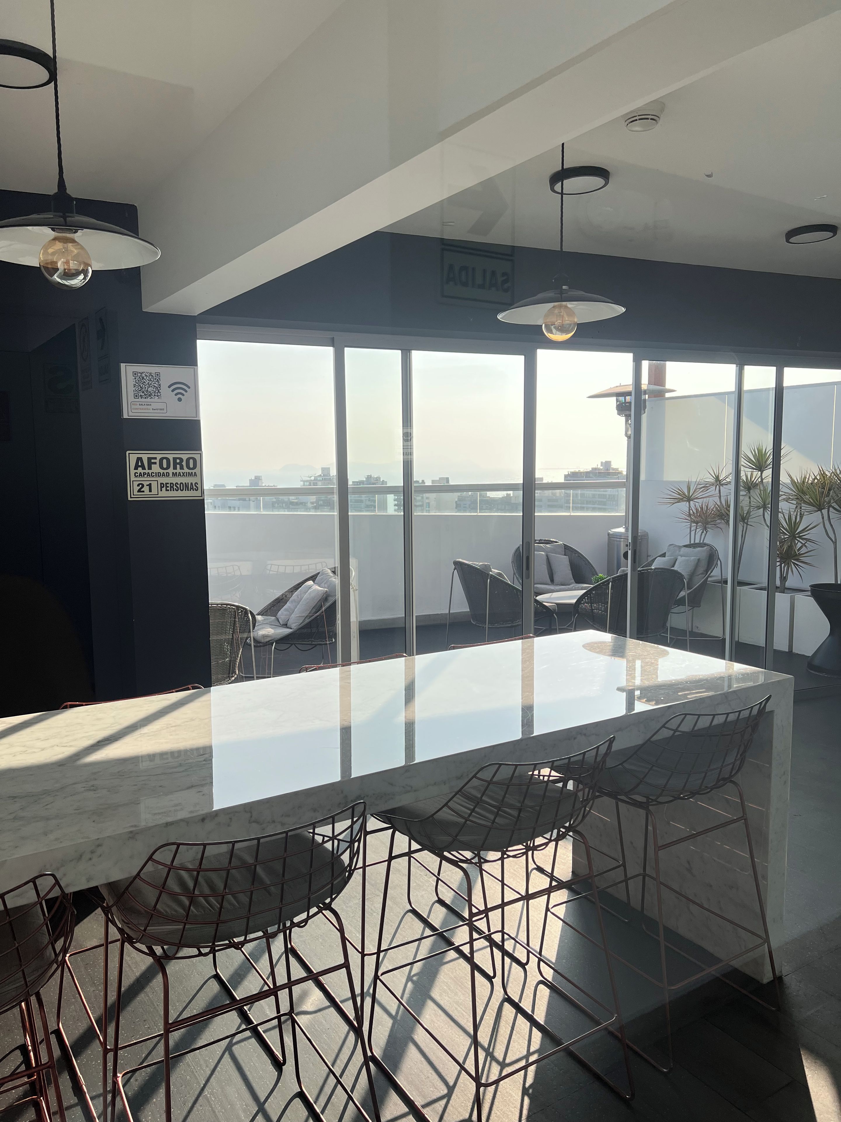 Un espace repas moderne avec une table en verre et des chaises en métal, de grandes fenêtres offrant une vue sur l’eau, et un panneau indiquant une capacité maximale de 21 personnes.