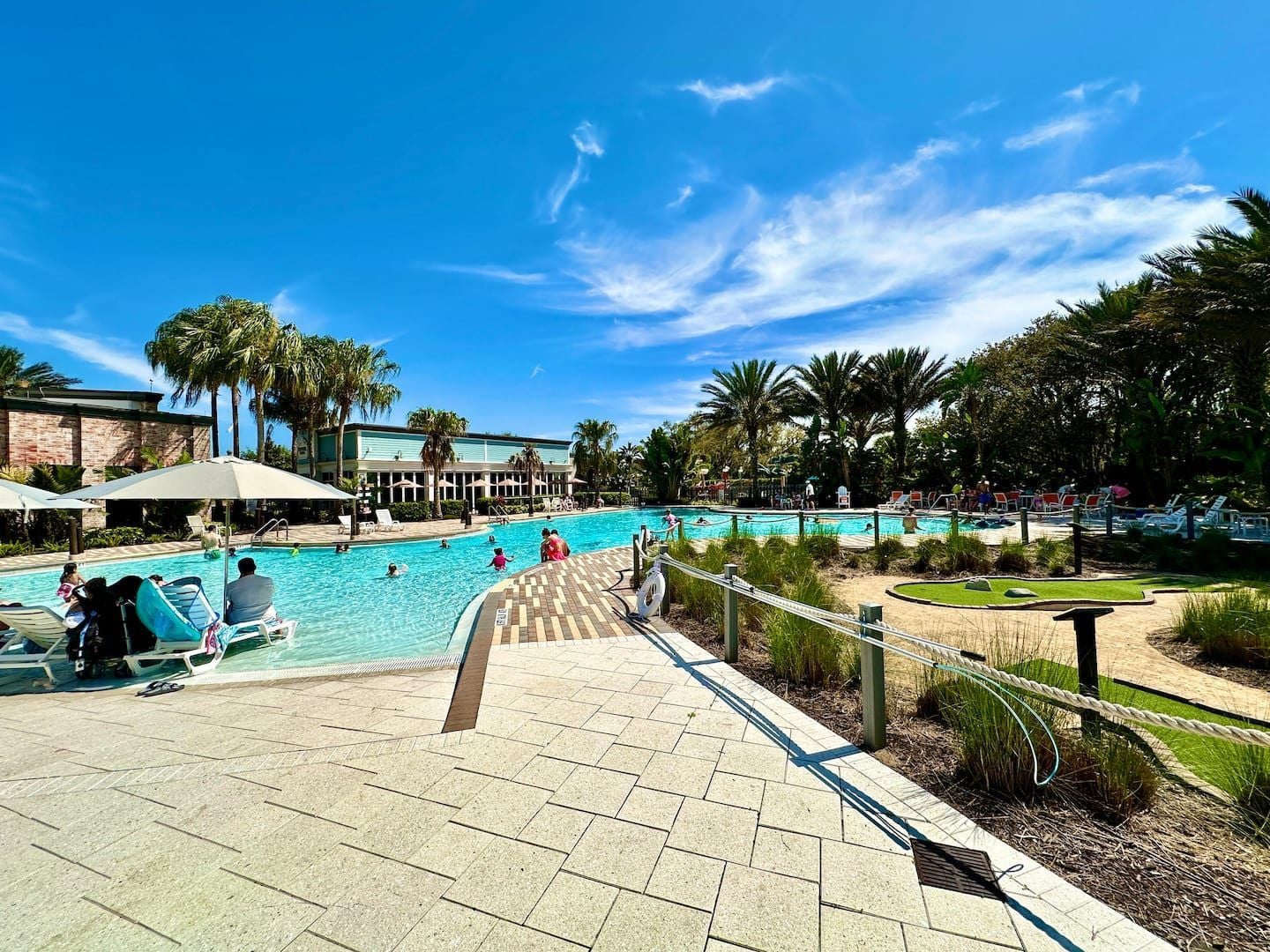 A swimming pool area with lounge chairs and umbrellas, surrounded by palm trees and a building in the background.