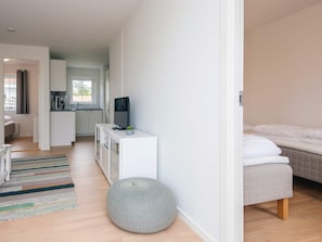 A modern, well-lit room with a bed, a television on a white stand, a kitchen area in the background, and a striped rug on the floor. - 4 Star Holiday Home in Laeso (Læsø)