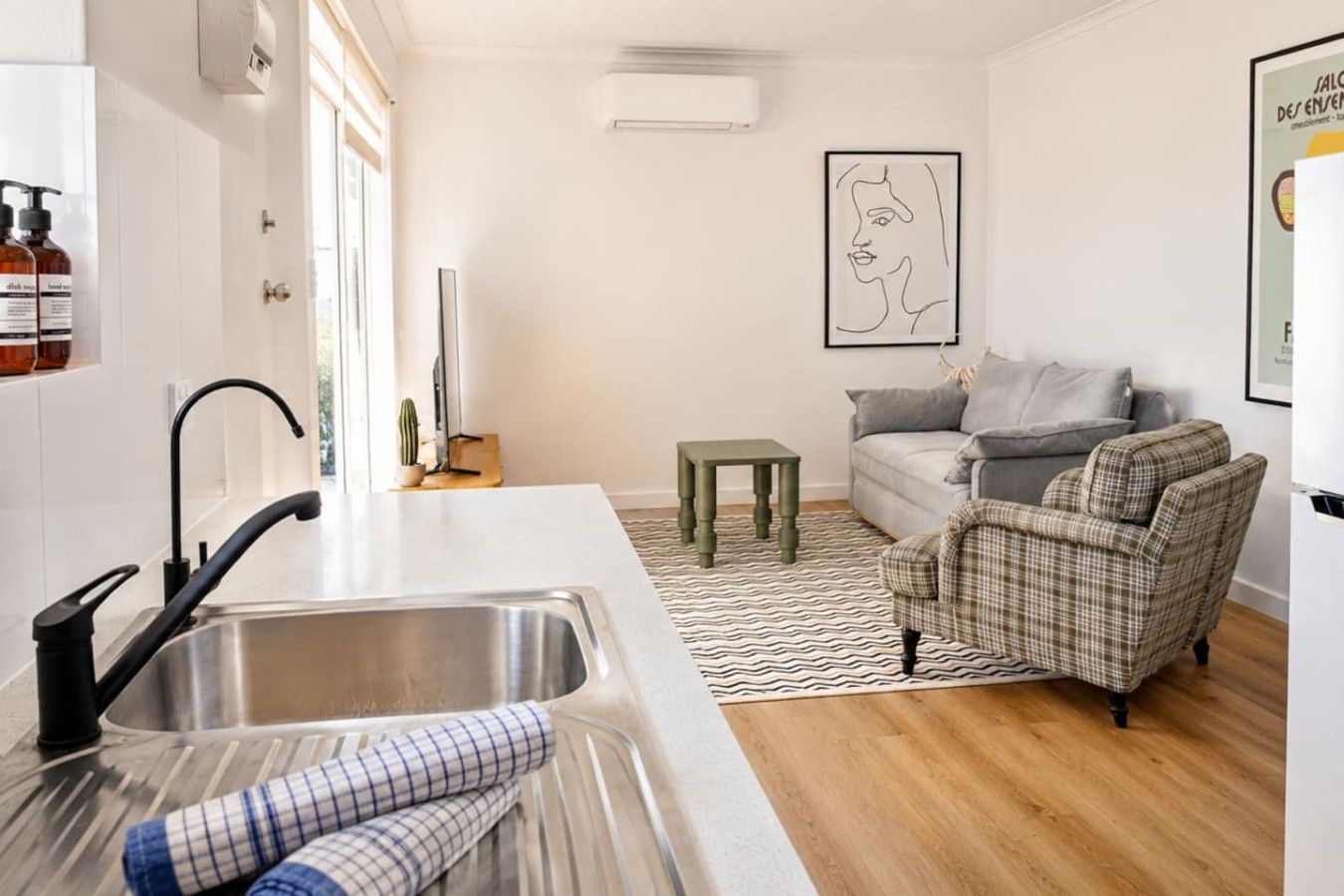 A modern kitchen with a stainless steel sink, a small table, and a grey sofa in a living area.