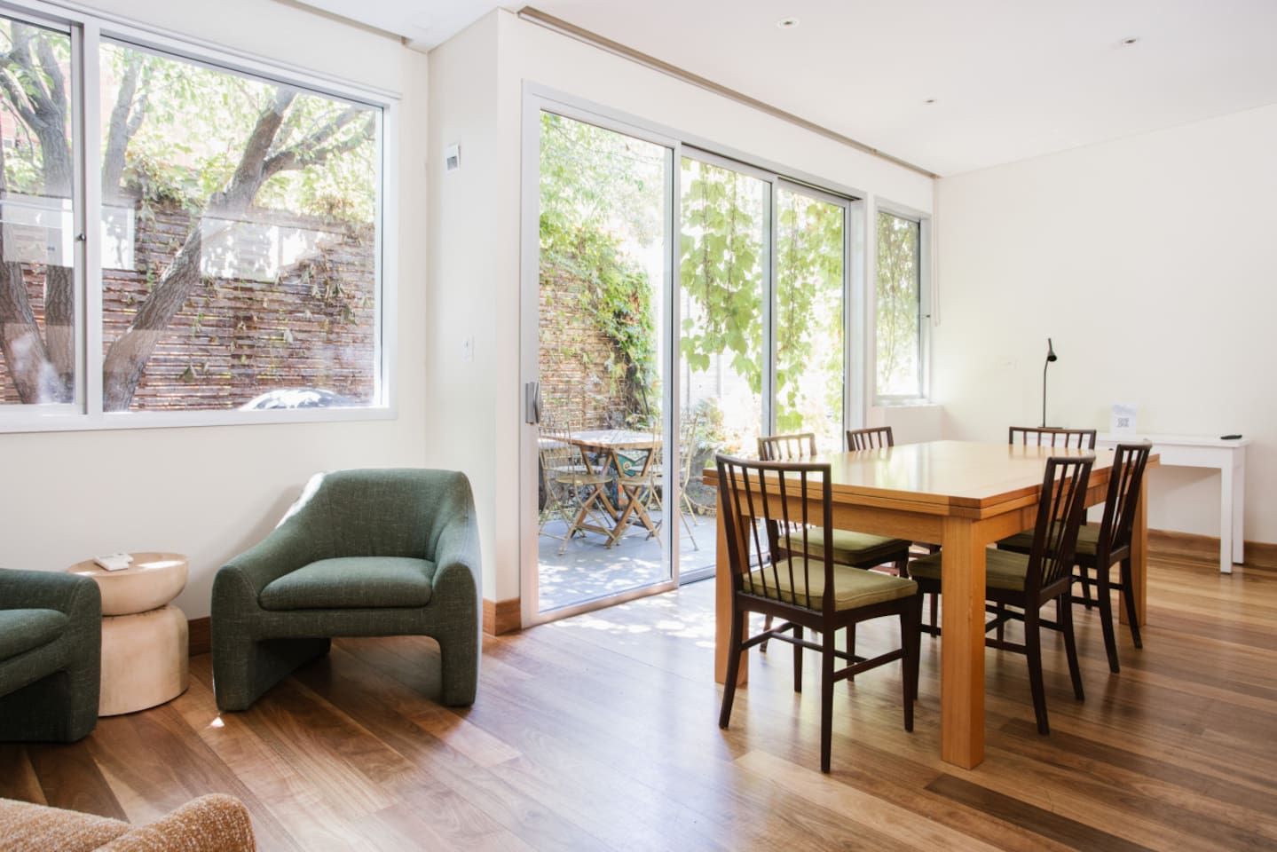 Un espace de restauration moderne avec une table et des chaises en bois, de grandes fenêtres et une vue sur un mur de briques et la verdure à l’extérieur.