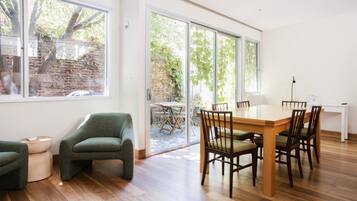 A modern dining area with a wooden table and chairs, large windows, and a view of a brick wall and greenery outside.