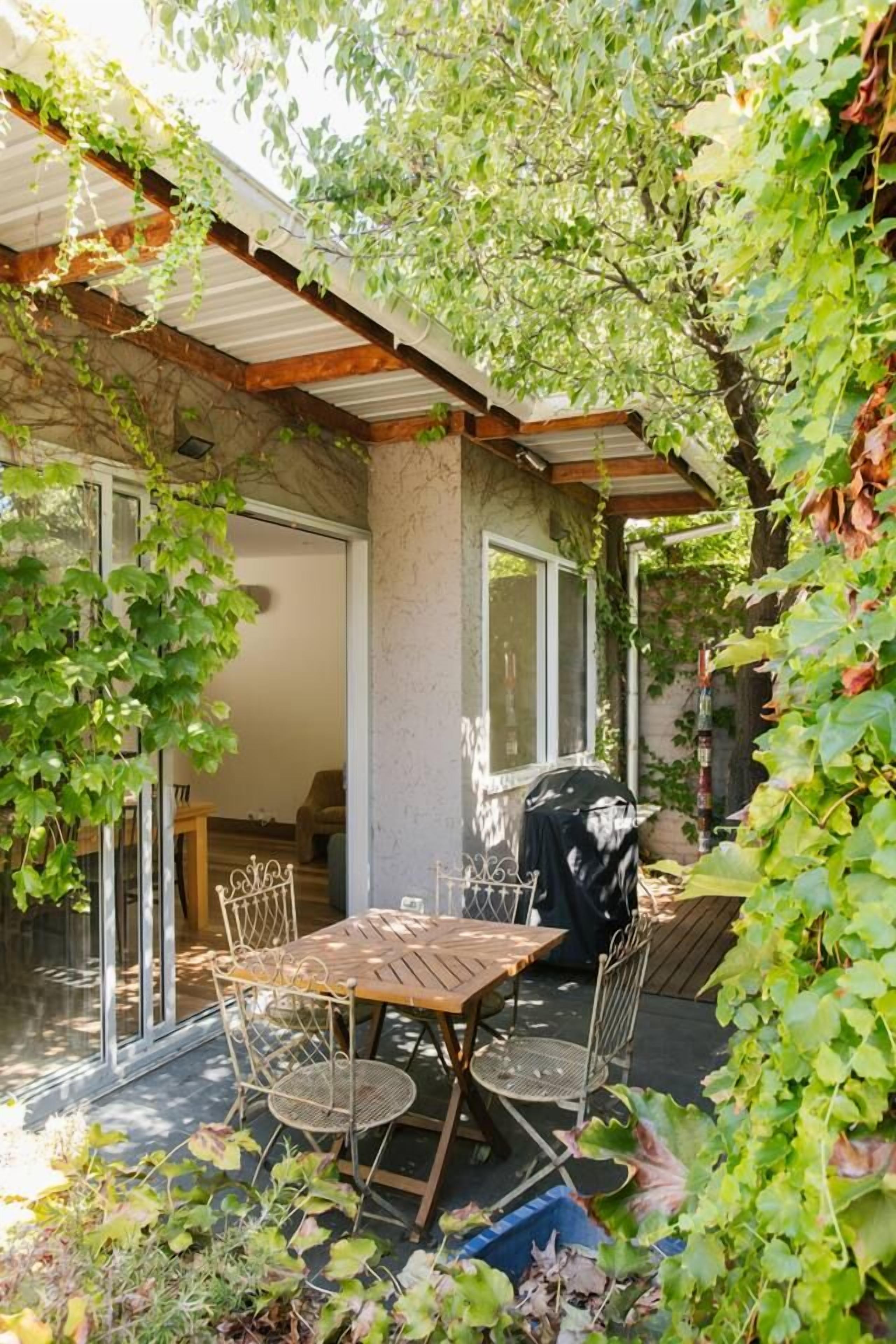 Une terrasse avec une table et des chaises en bois, un barbecue et un espace couvert donnant sur l’intérieur.