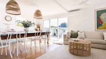 Een moderne woonkamer met een bank, een eethoek met stoelen, een groot raam en een plafondventilator.