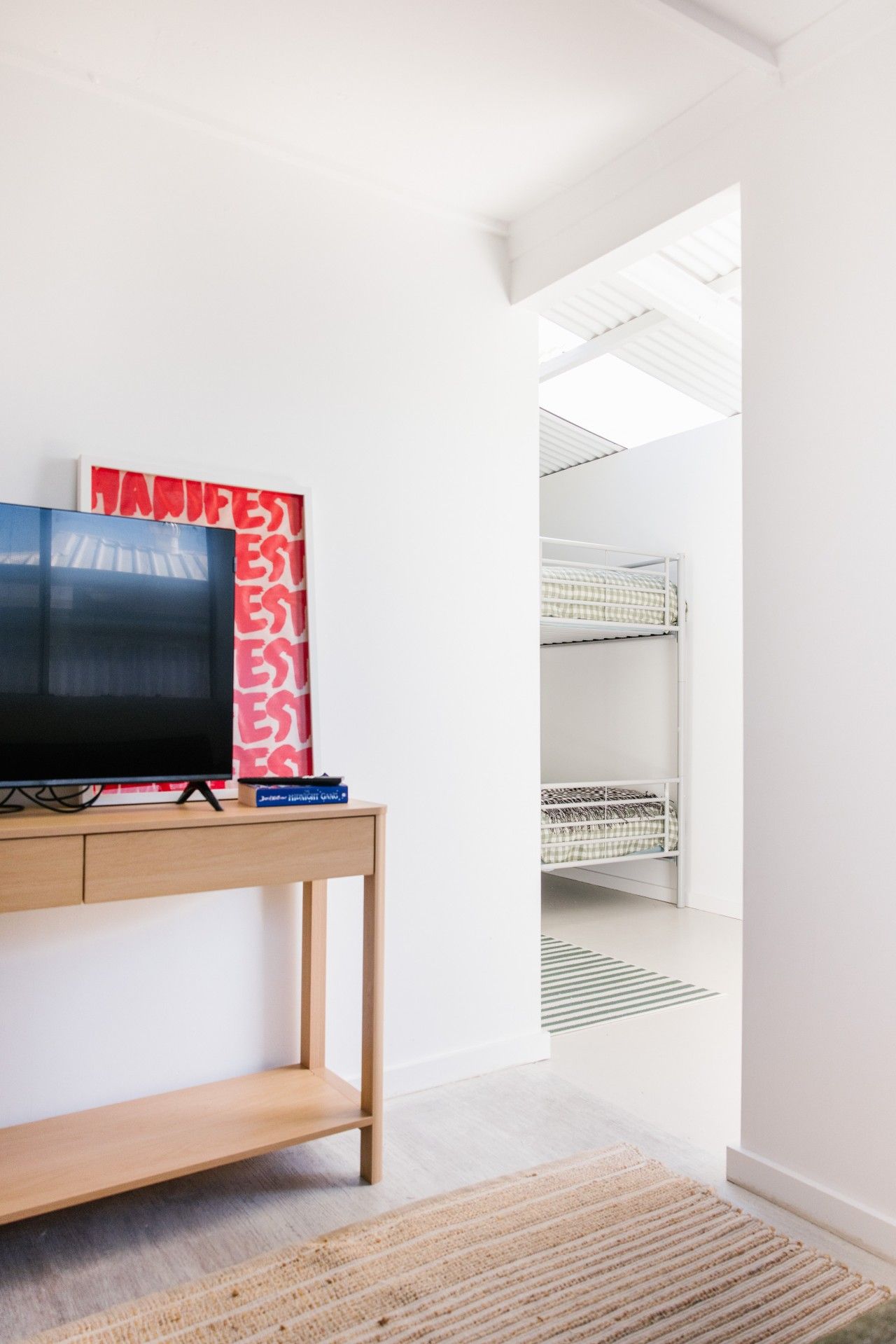 Quarto minimalista com televisão sobre um console de madeira, um objeto emoldurado em vermelho e branco na parede, e uma cama dupla ao fundo.
