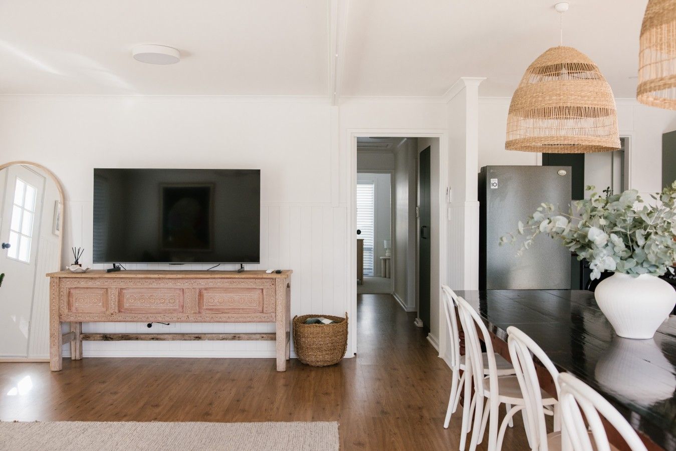 A modern kitchen with a large flat-screen TV, a wooden cabinet, a refrigerator, and a dining area with white chairs.