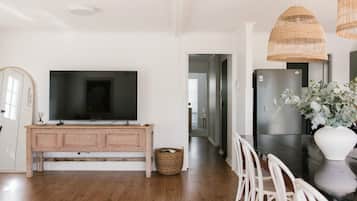 A modern kitchen with a large flat-screen TV, a wooden cabinet, a refrigerator, and a dining area with white chairs.