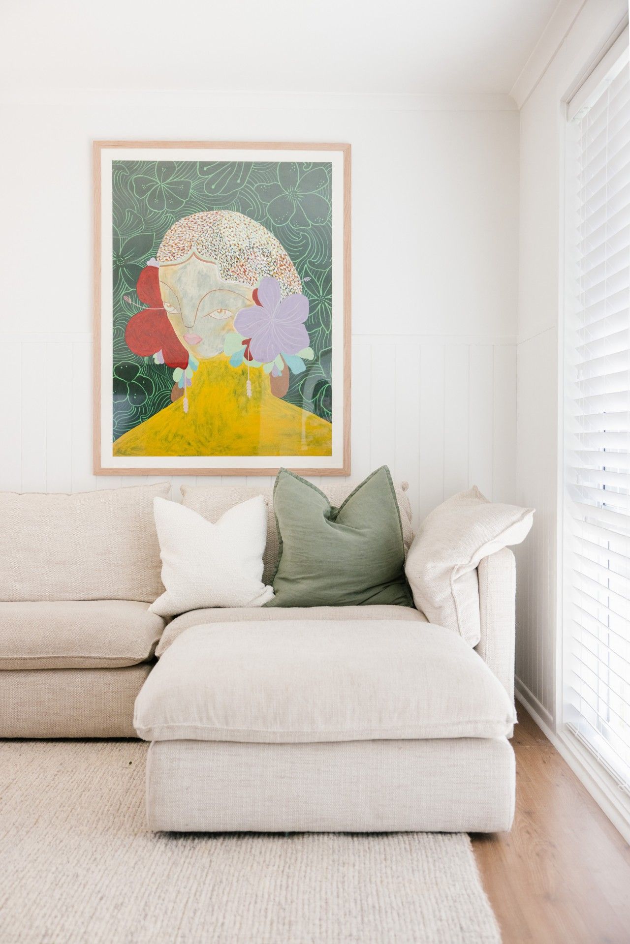 A living room with a beige sofa, a framed abstract painting, and a window with blinds.