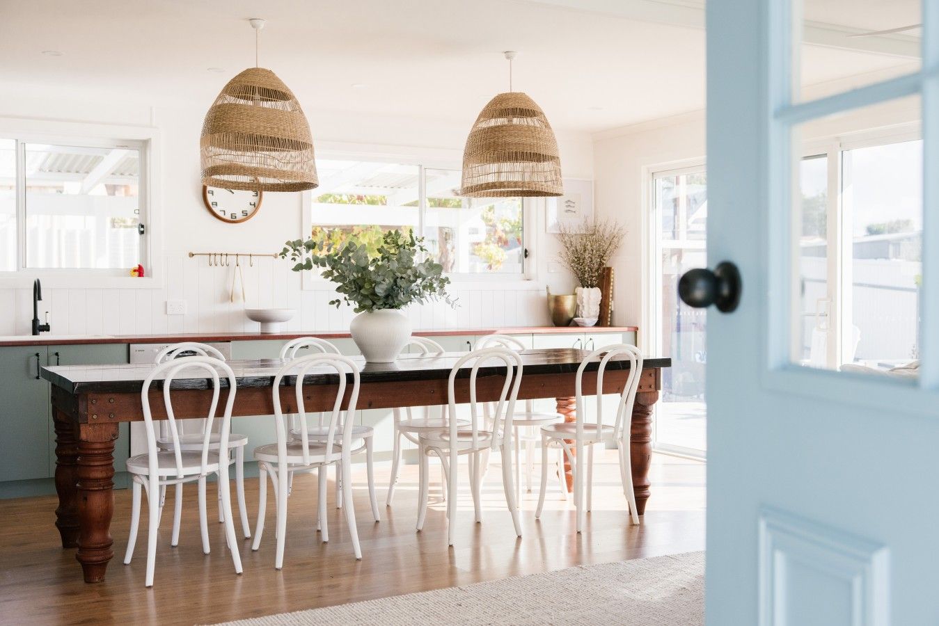 A kitchen with a wooden island, white chairs, hanging wicker pendant lights, and a potted plant.