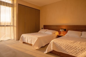 Una camera d'albergo con due letti, un armadio in legno, un comodino con lampada e una finestra con tende trasparenti.