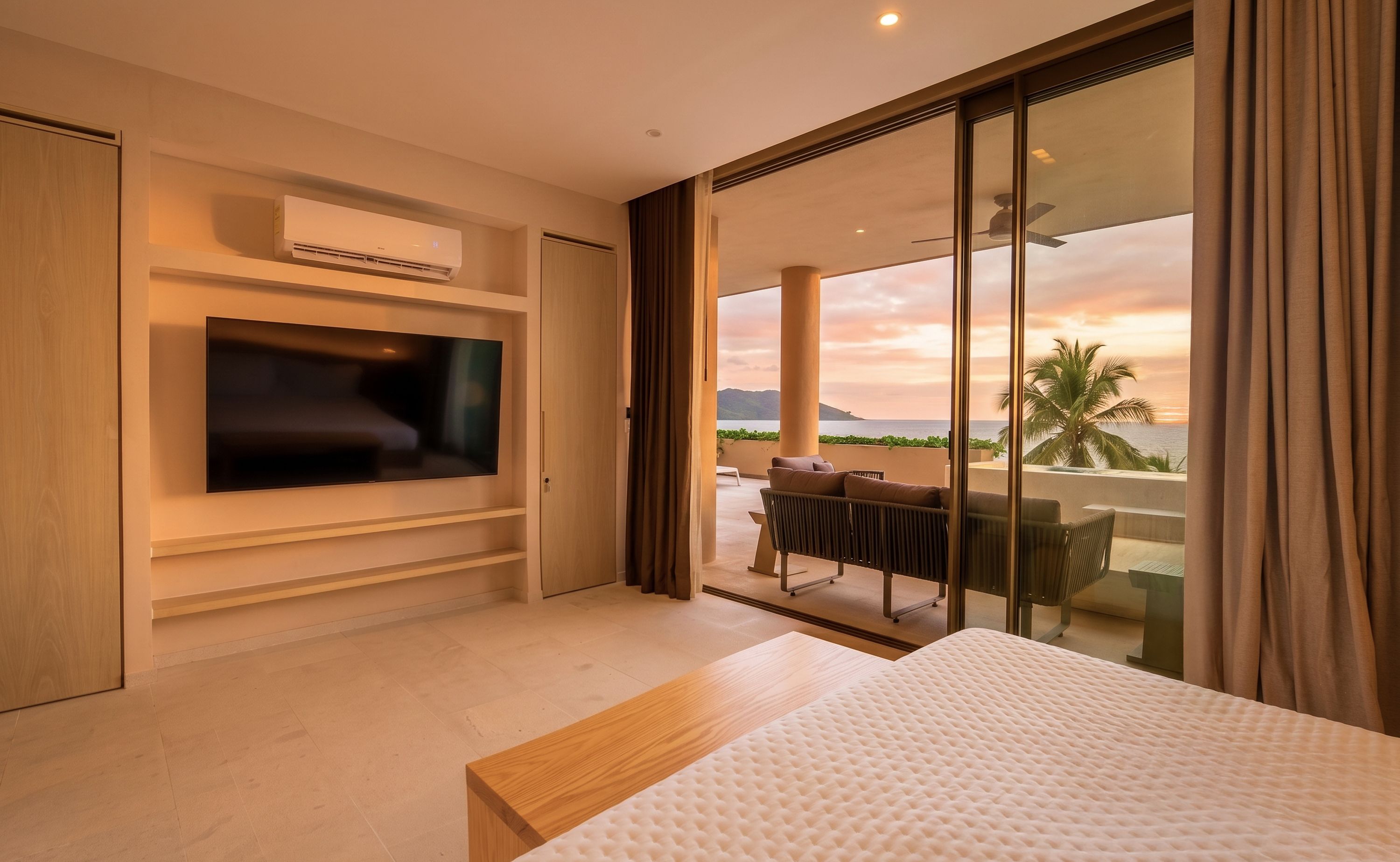 A modern hotel room with a flat-screen TV, a bed, a balcony with a view of the sea, and a ceiling fan.