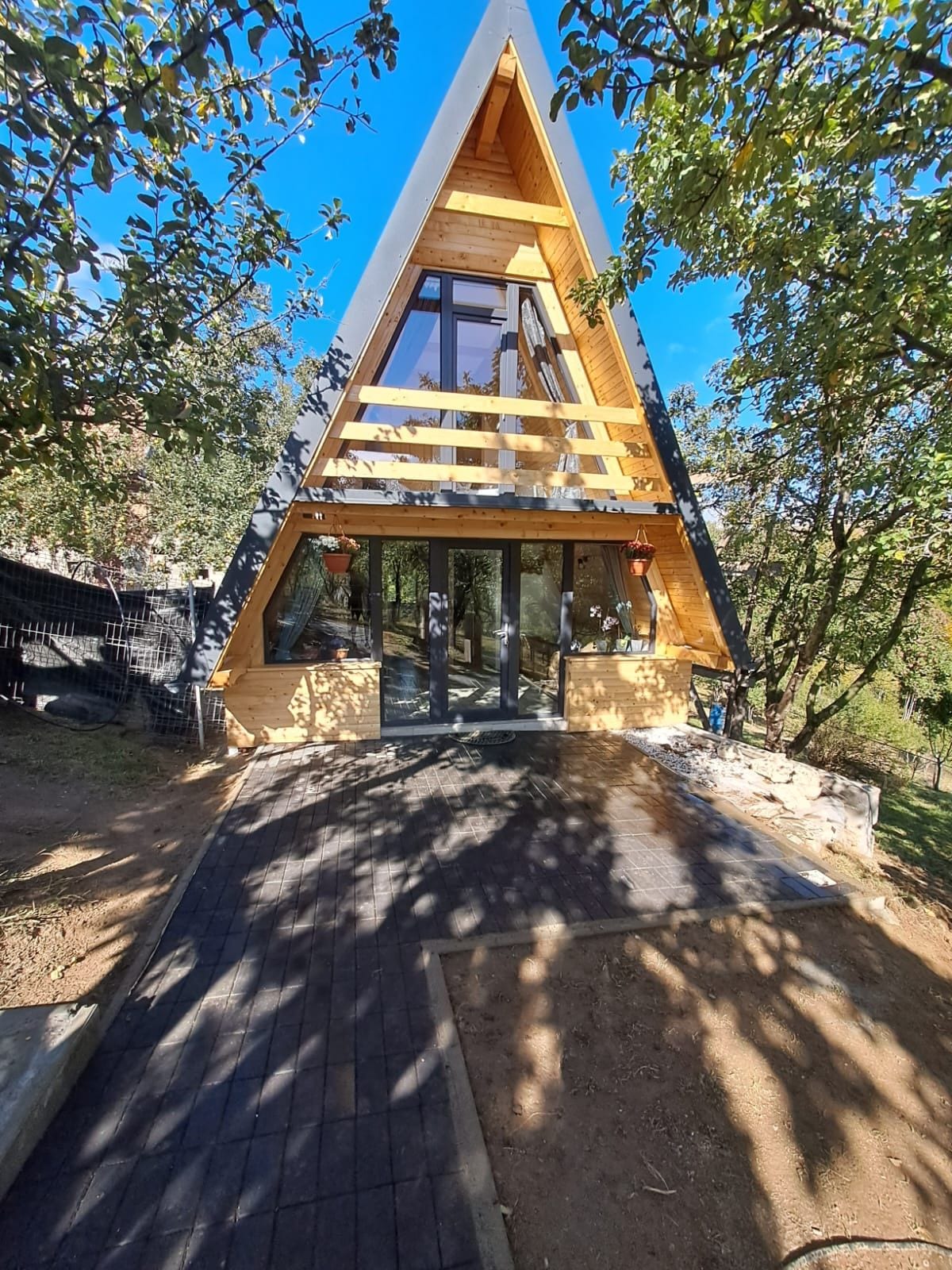 Une structure triangulaire en bois, dotée d’une entrée en verre et entourée d’arbres.