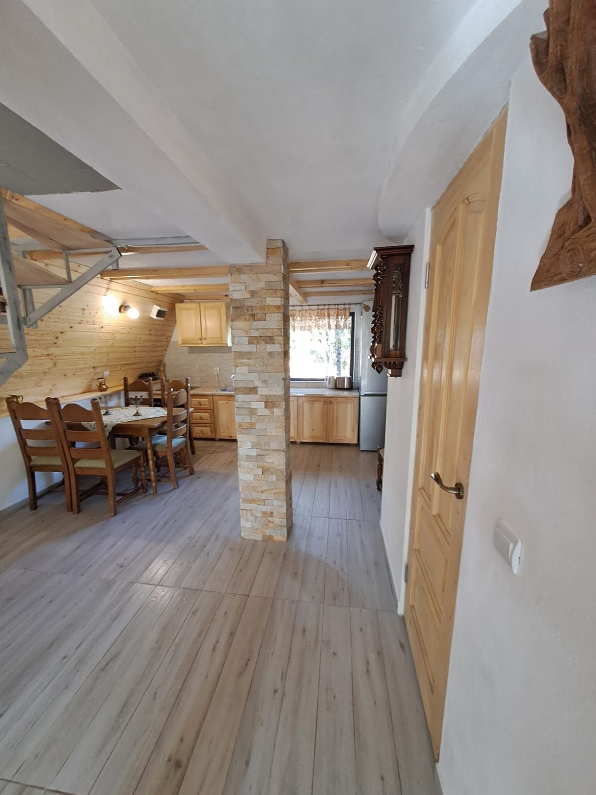 Une table à manger en bois avec des chaises, un pilier en pierre et un coin cuisine avec des placards en bois.