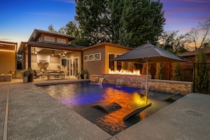 Ein Garten mit Schwimmbecken, Feuerstelle und überdachter Terrasse.