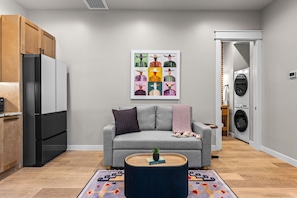 Ein modernes Wohnzimmer mit einem grauen Sofa, einem Couchtisch, einem Wandbild und einer Waschmaschine im Hintergrund.