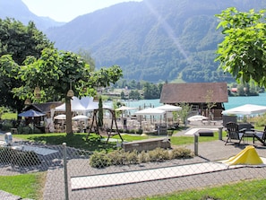 Un espace extérieur aménagé au bord du lac, avec des tables et des parasols, entouré de verdure et offrant une vue sur les montagnes en arrière-plan.