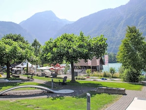 Un parc a la vora d'un llac amb taules de pícnic, un espai de jocs i un edifici amb teulada vermella.