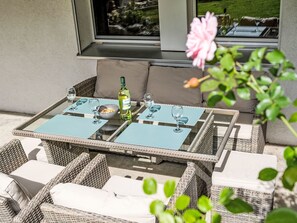 An outdoor seating area with wicker furniture and glass-top tables, a bottle of wine, and a view of a garden through a window. - Ferienwohnung Enzian by Interhome (Lungern)