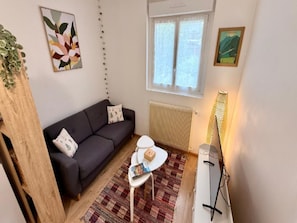 A compact living space with a dark sofa, a small round table, a radiator, and a window with curtains. - Family Apartment in Mont-dore (Mont-Dore)