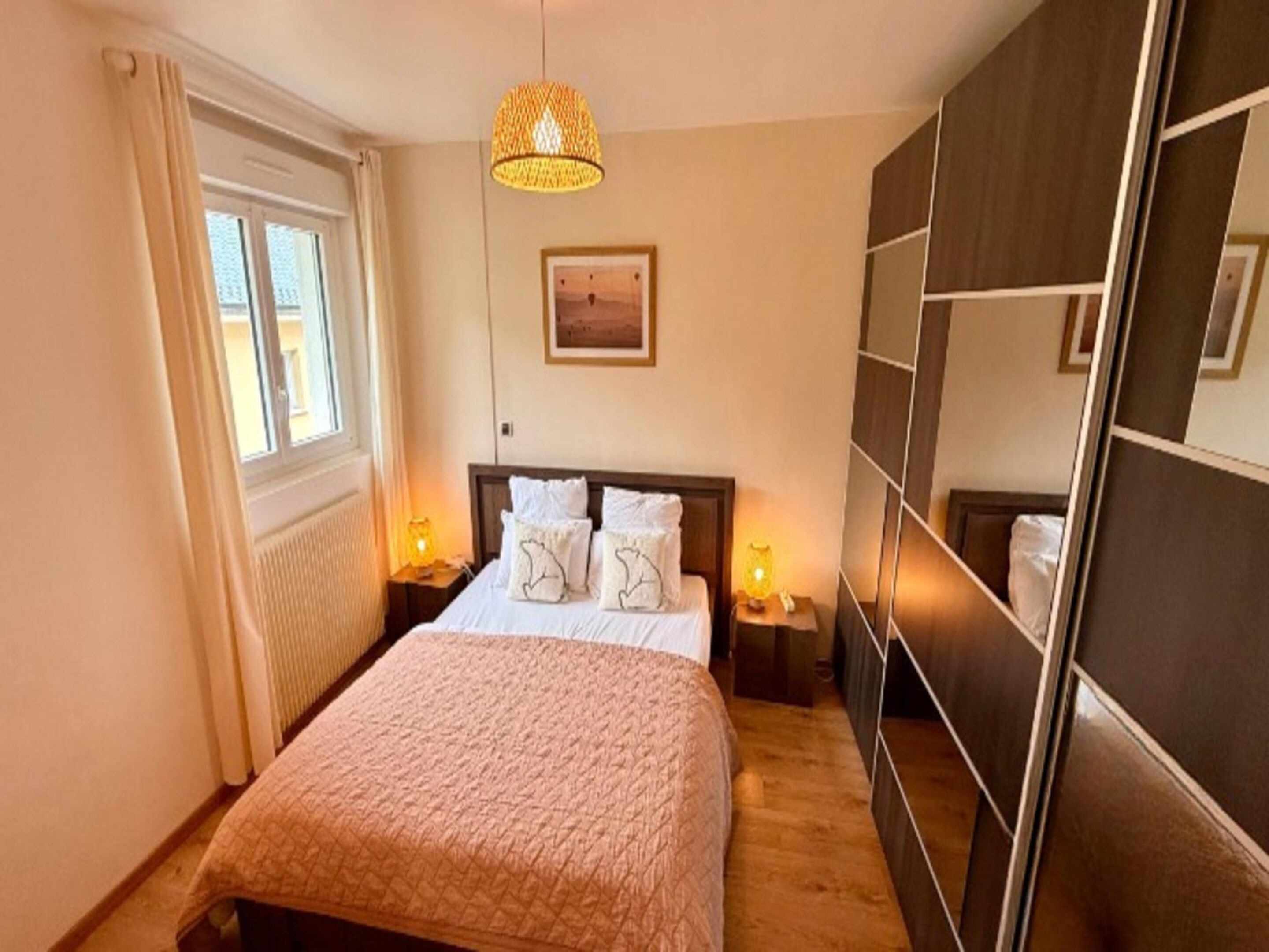 A bedroom with a bed, bedside lamps, a window, a wall-mounted artwork, and a mirror.