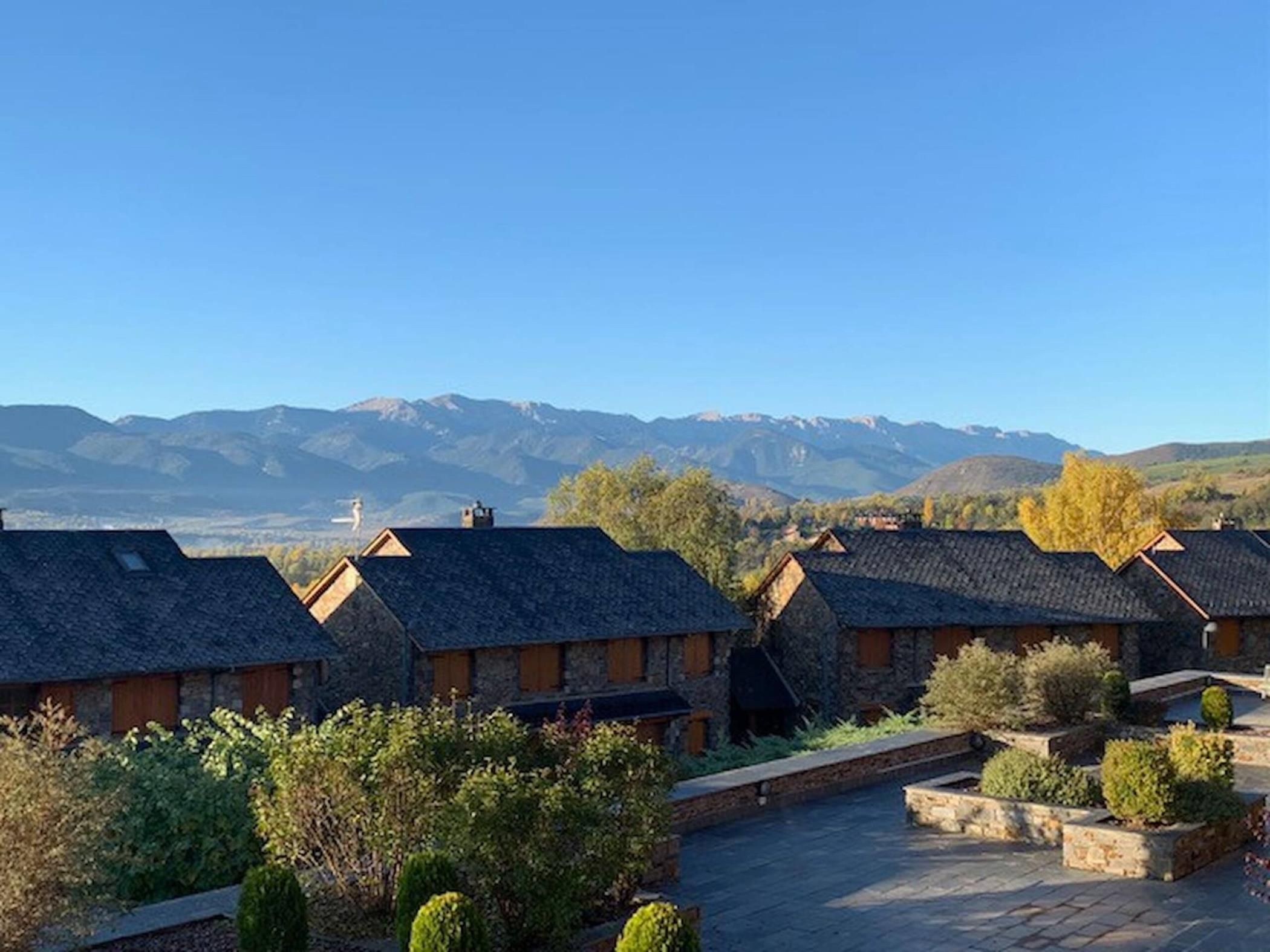 A row of stone buildings with sloped roofs, surrounded by greenery and a mountainous backdrop.