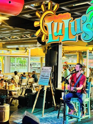 Een muzikant treedt op in een locatie met een bord waarop 'Lucy Buffett's' staat en een bord dat fooien aanmoedigt voor 'Logan Lassitter' via CashApp.