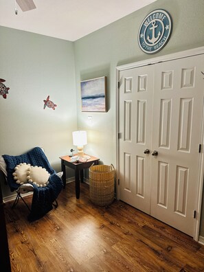 Una habitación con piso de madera, una silla con una manta azul, una lámpara, una cesta de mimbre, un cuadro y una puerta con un cartel de bienvenida.