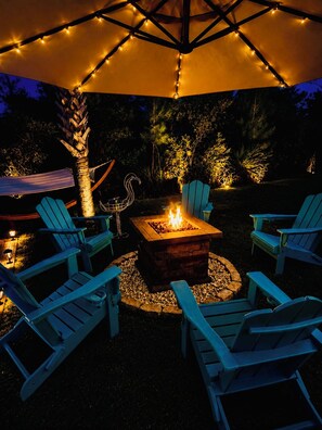 An outdoor seating area with a fire pit, surrounded by Adirondack chairs, under a canopy with string lights. - Resting Beach Face Sleeps 10, 2 Kings, Bunks, Mini Bar, 2 Pools, Beach 6 Blocks (Gulf Shores)